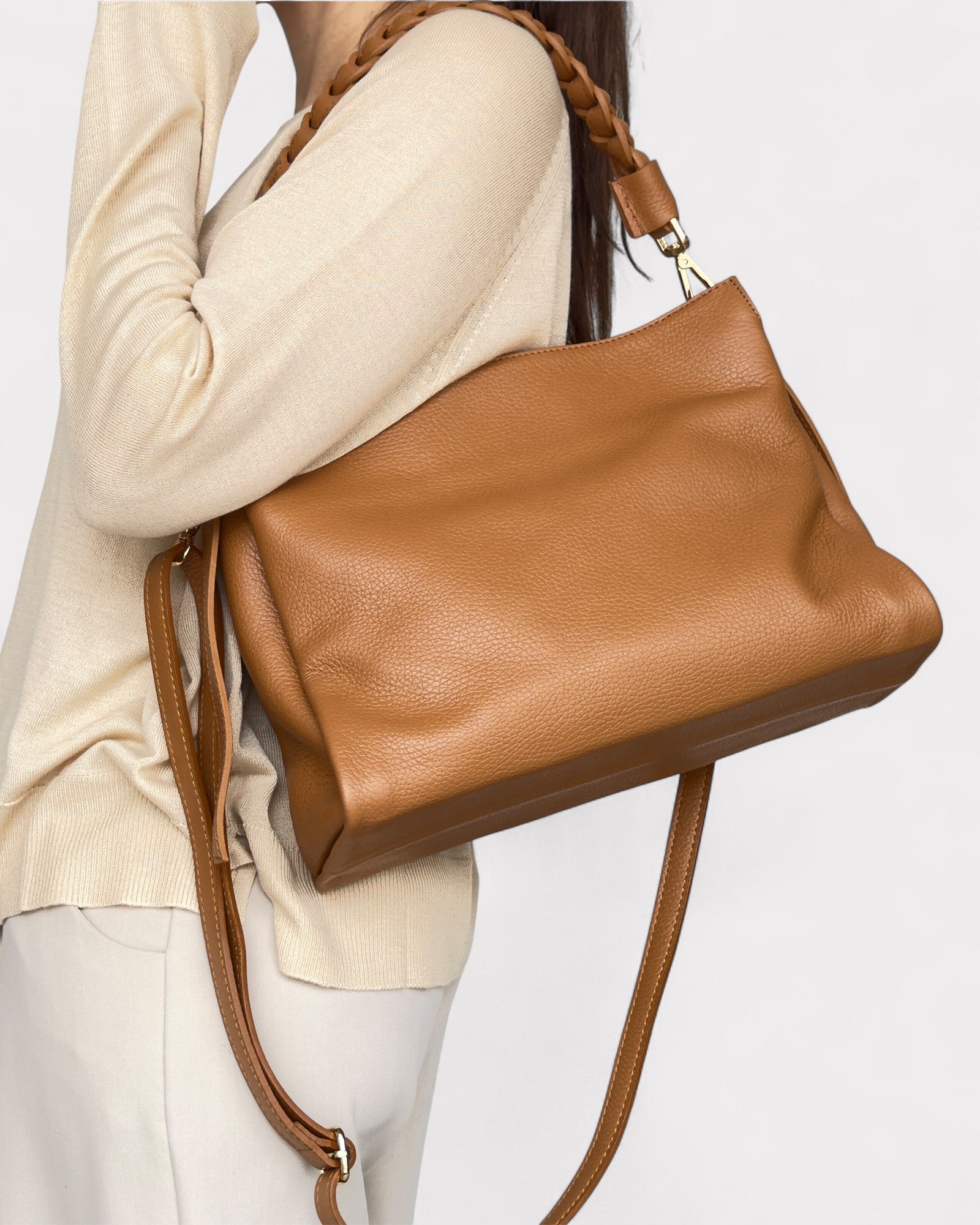 Tan leather handbag being carried by a person wearing a beige sweater and white pants on a light gray background