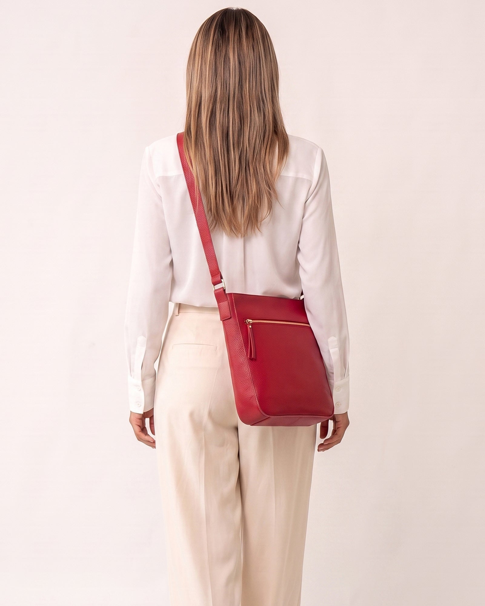 A woman viewed from behind wearing a white blouse and cream trousers, carrying a red leather crossbody bag with a gold zipper pull and gold hardware against a plain off-white background.