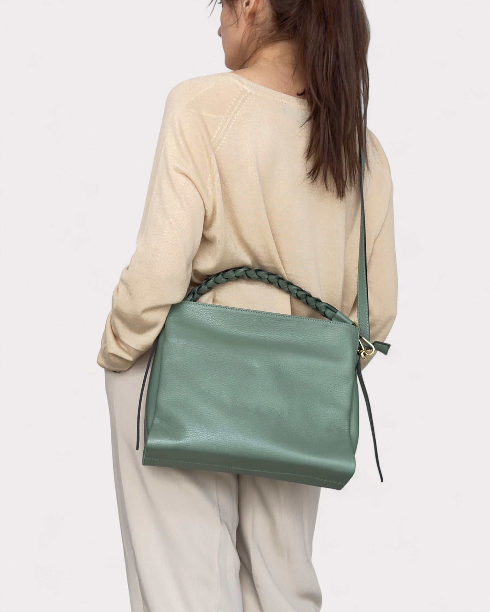 Person wearing a light beige sweater and light-colored pants with a green handbag over one shoulder on a white background