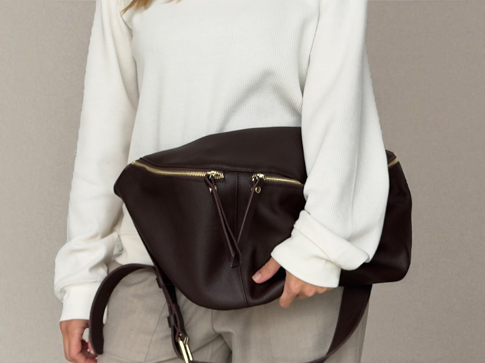 Person holding a brown leather bag with gold zippers against a neutral background