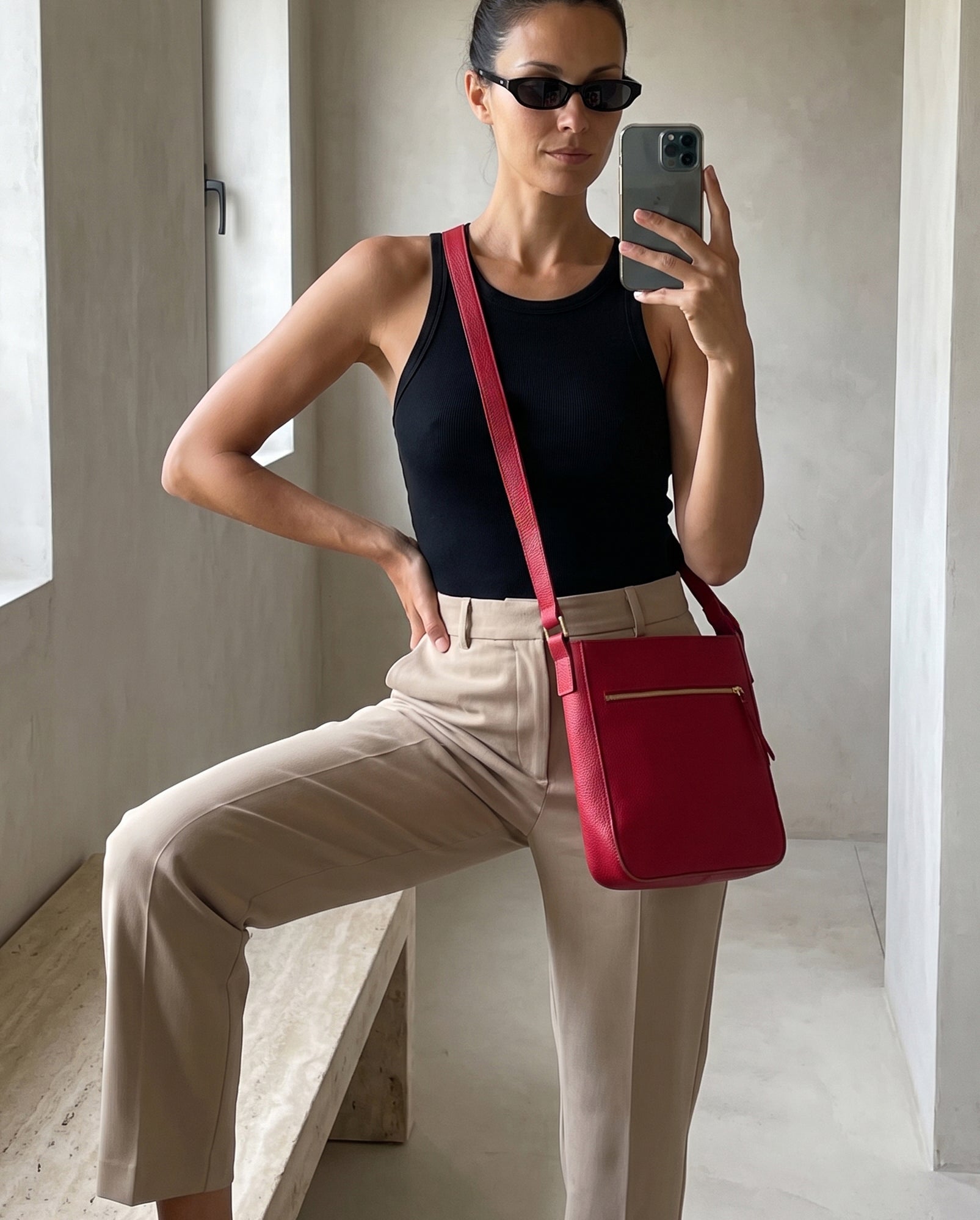 A woman takes a mirror selfie wearing a black tank top, beige trousers, and black sunglasses, carrying a bright red leather crossbody bag with a gold zipper.