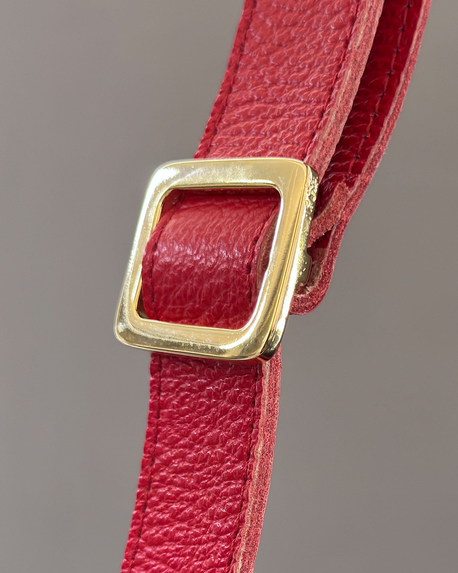 A close-up of a red textured leather strap with a square gold-tone metal buckle, set against a neutral gray background.