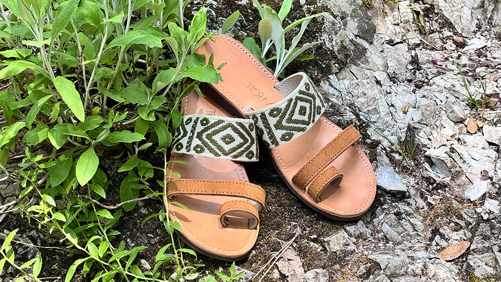 Pair of brown sandals with green patterned straps on a rocky surface with plants.