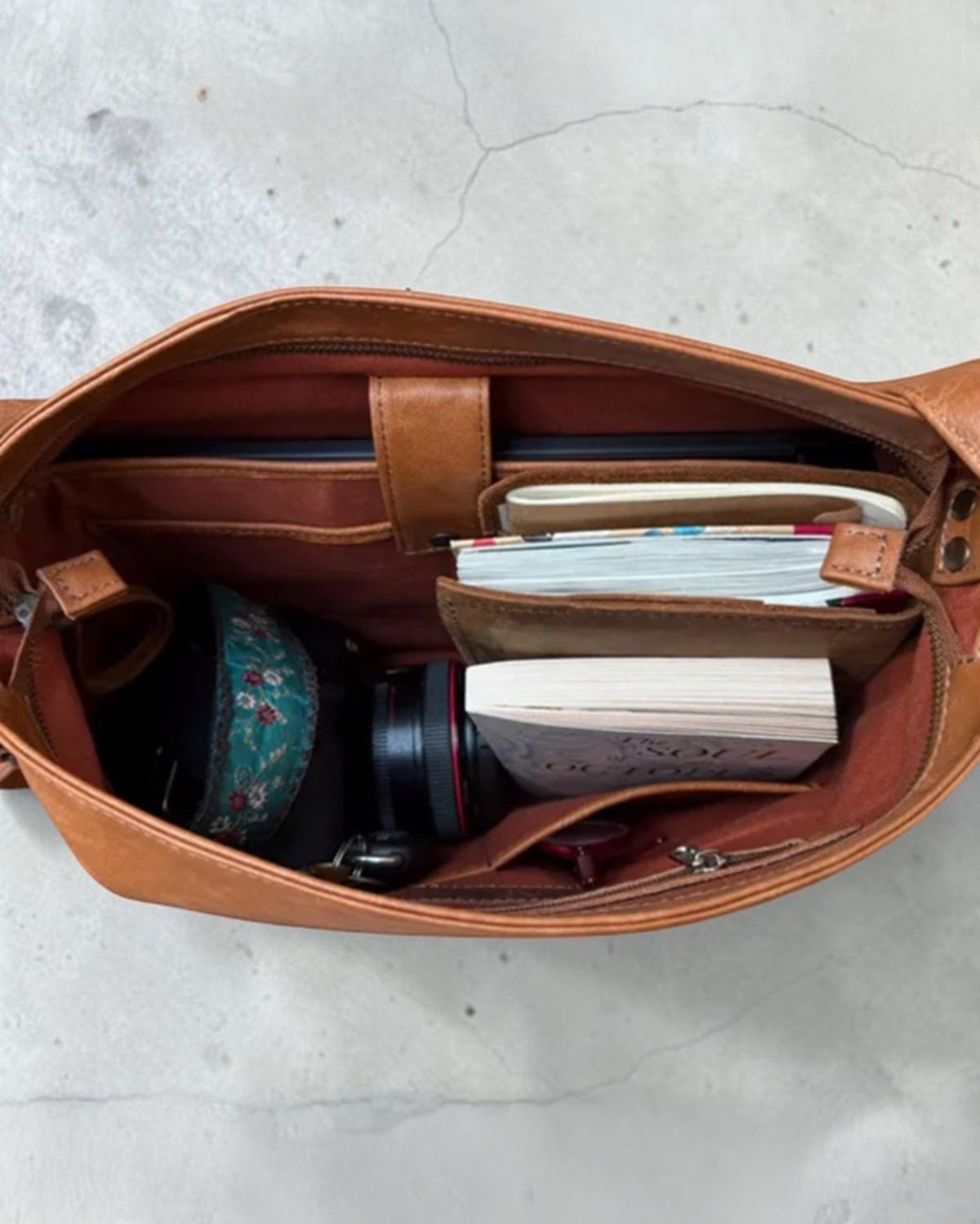 A tan leather convertible bag shown from above, filled with a wallet, notebook, camera lens, and other accessories, set against a light gray concrete floor.