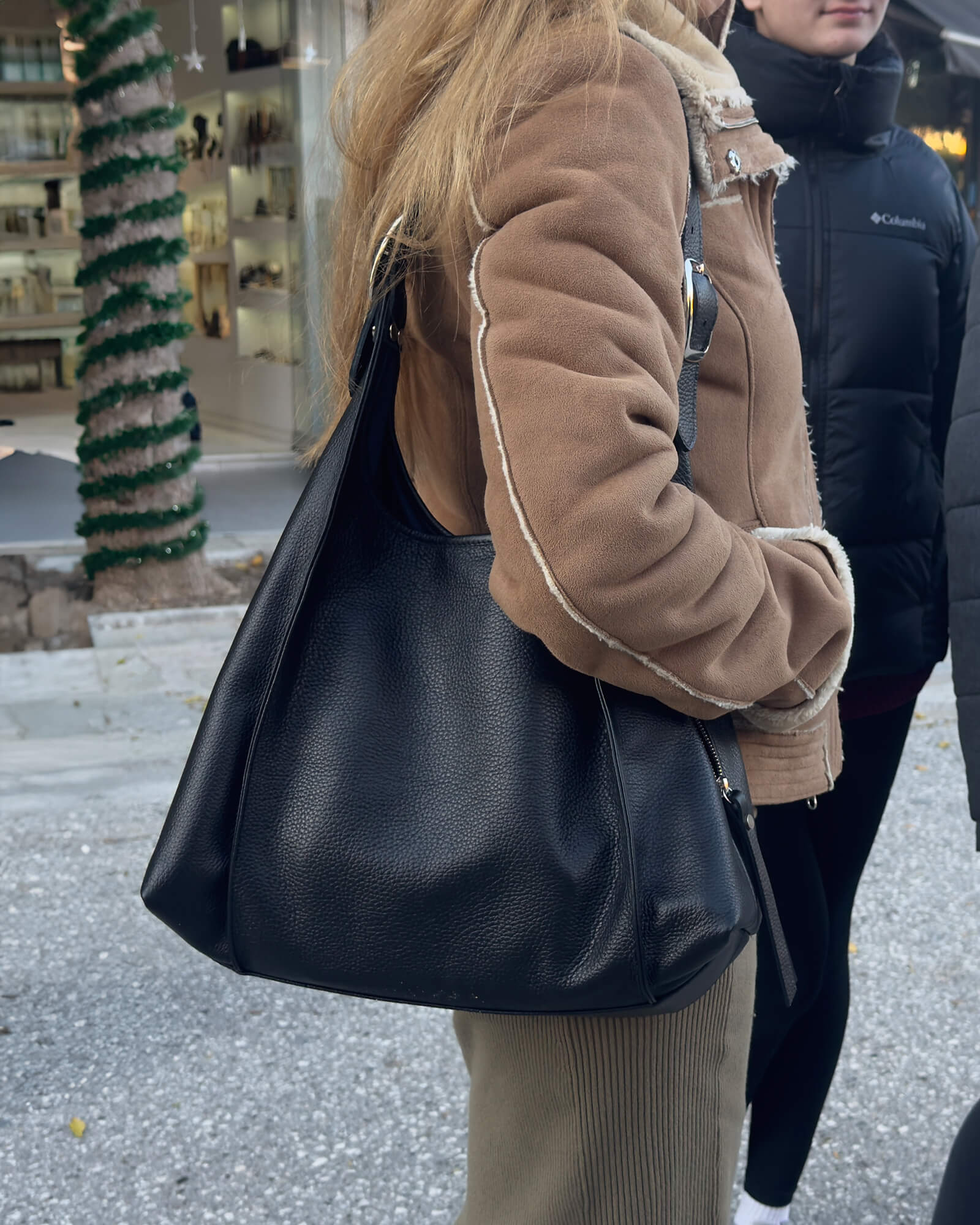 Person wearing a brown coat holding a black handbag on a street.