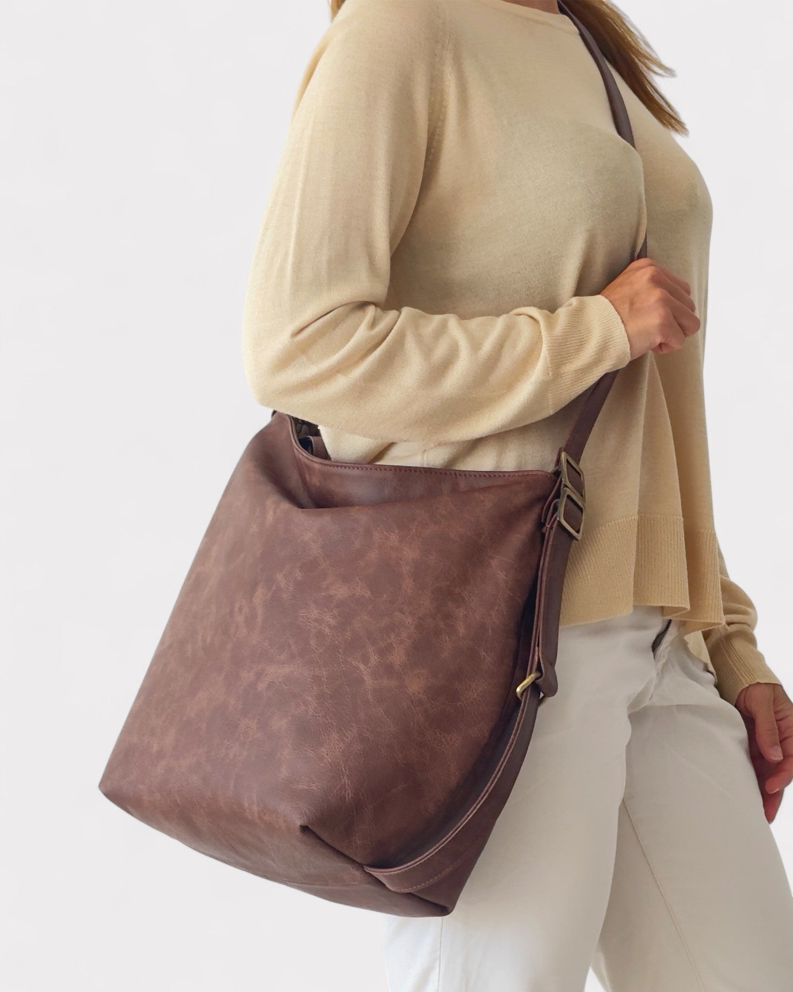Person wearing a beige sweater and white pants with a brown leather bag on a white background