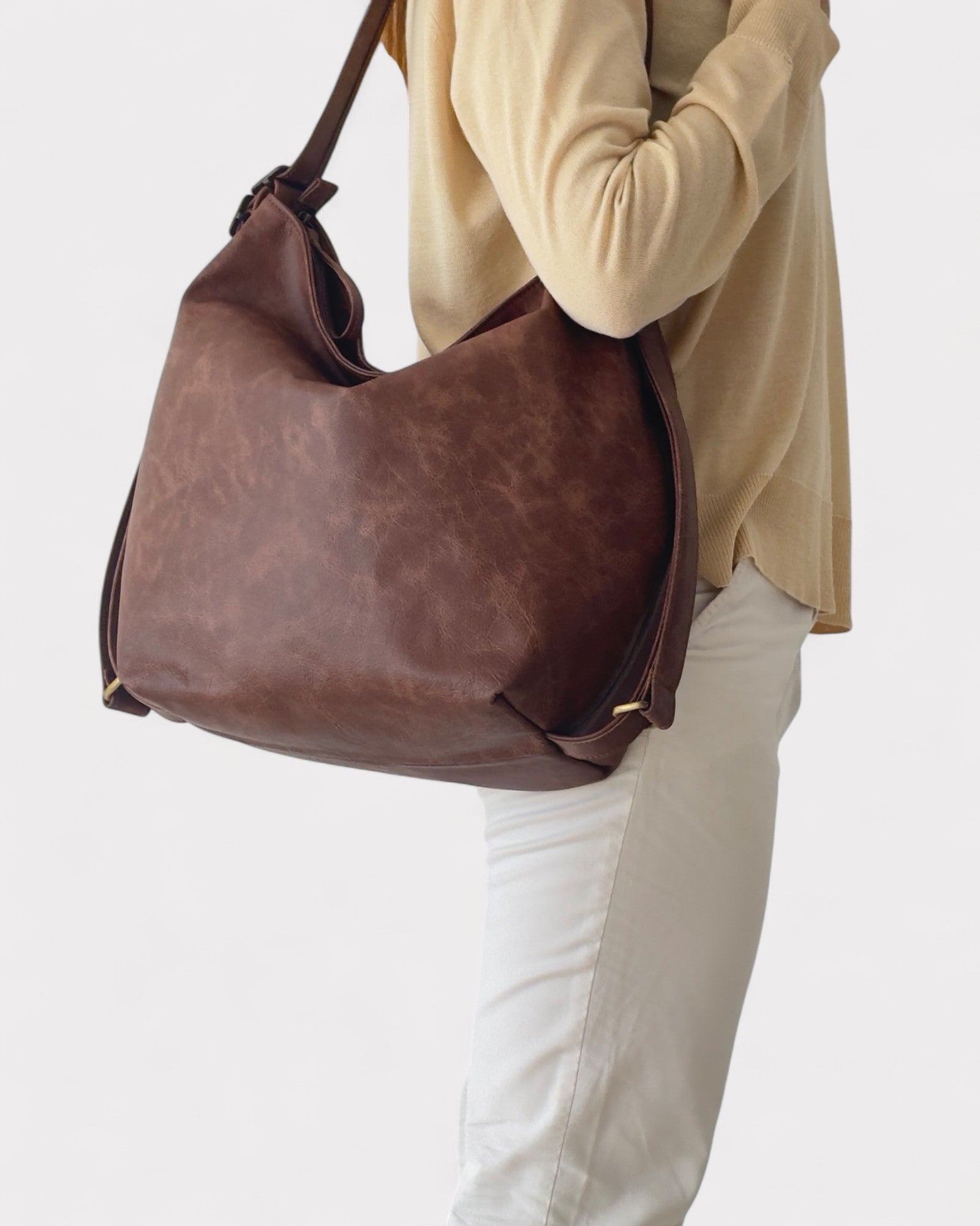 Brown leather handbag held by a person wearing a beige top and white pants on a light gray background
