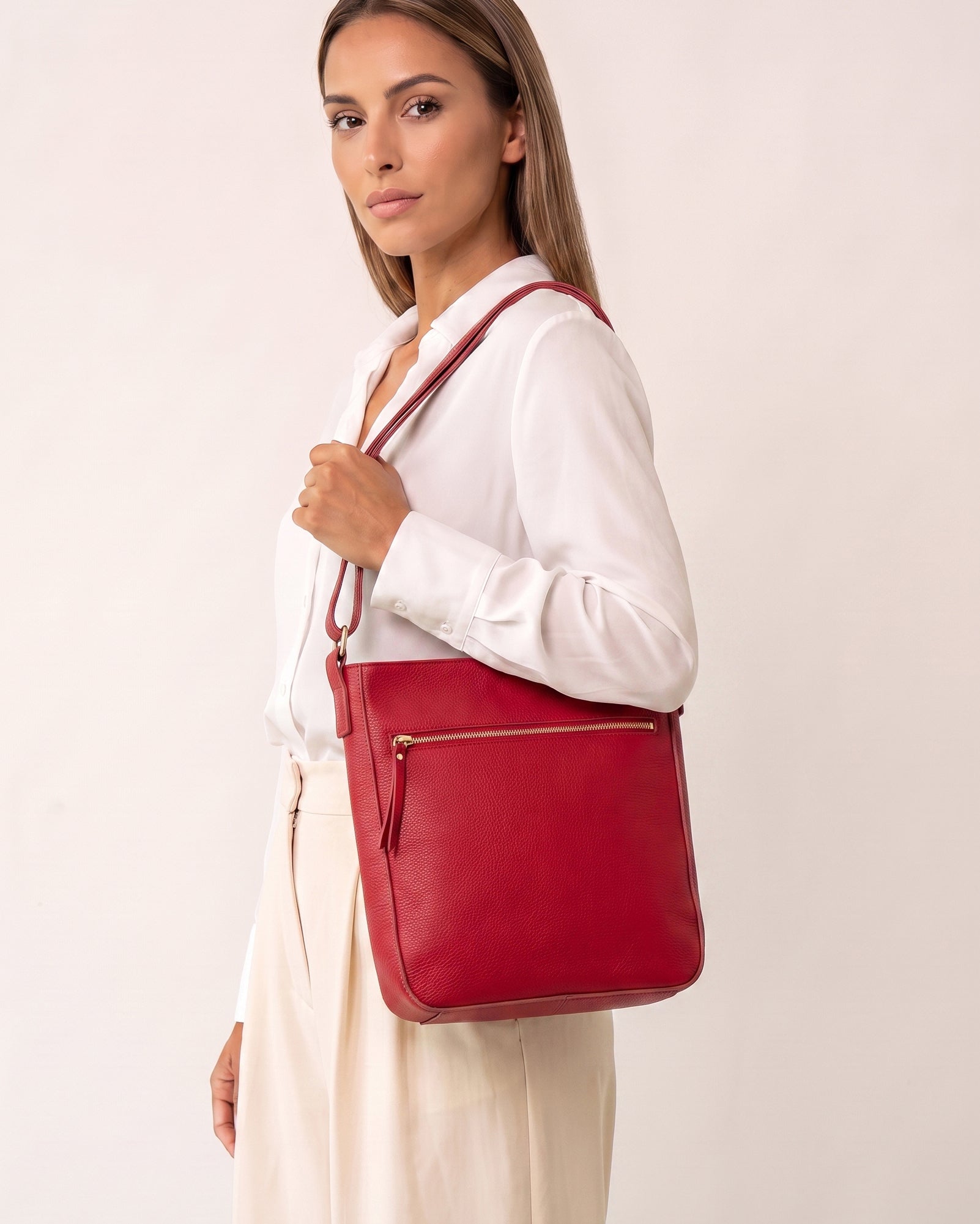 A woman wearing a white blouse and cream trousers carries a red leather shoulder bag with a matching strap against a plain off-white studio background.